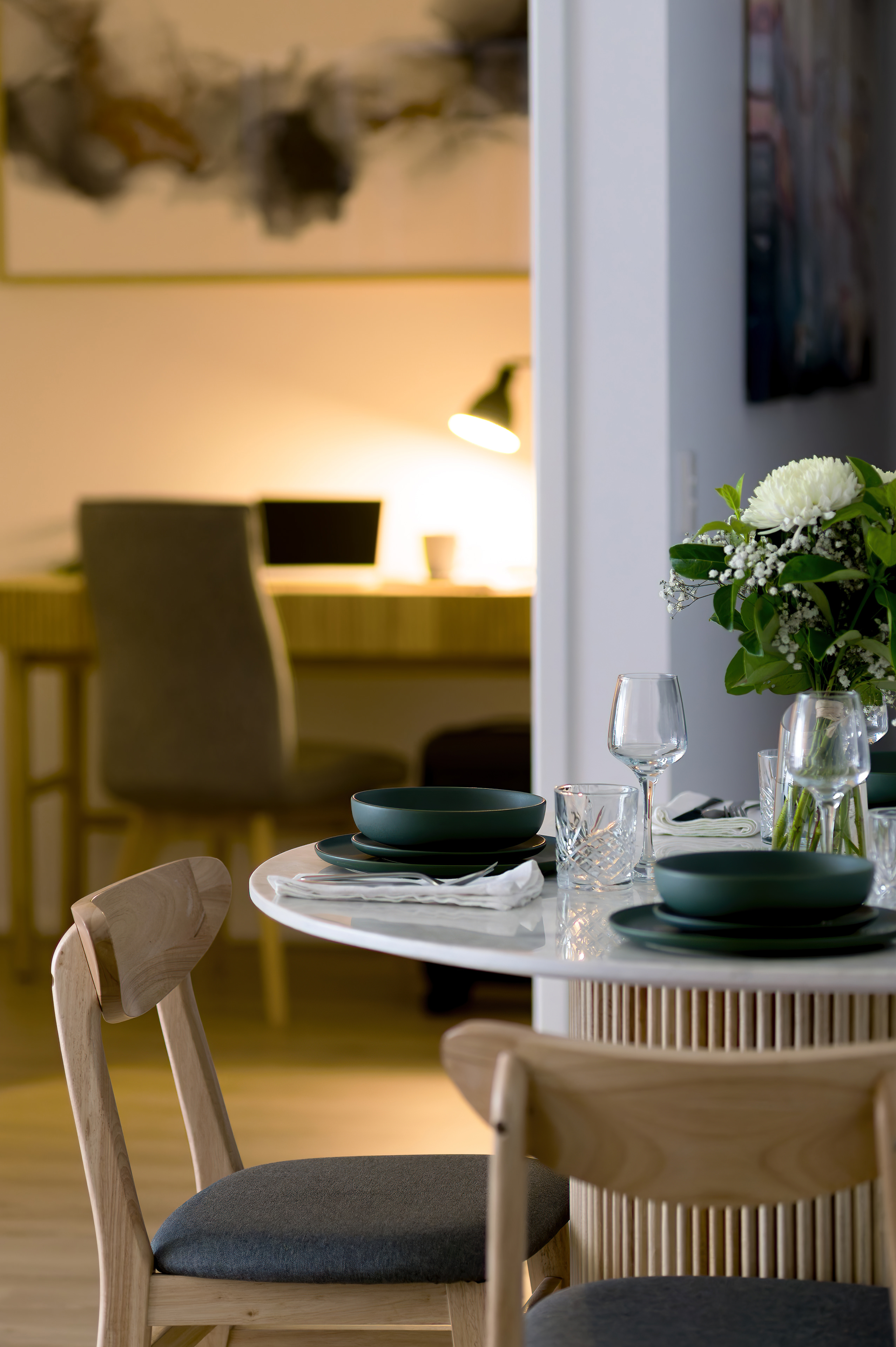 Apartment dining table set for dinner with a desk and workspace lit by a lamp behind